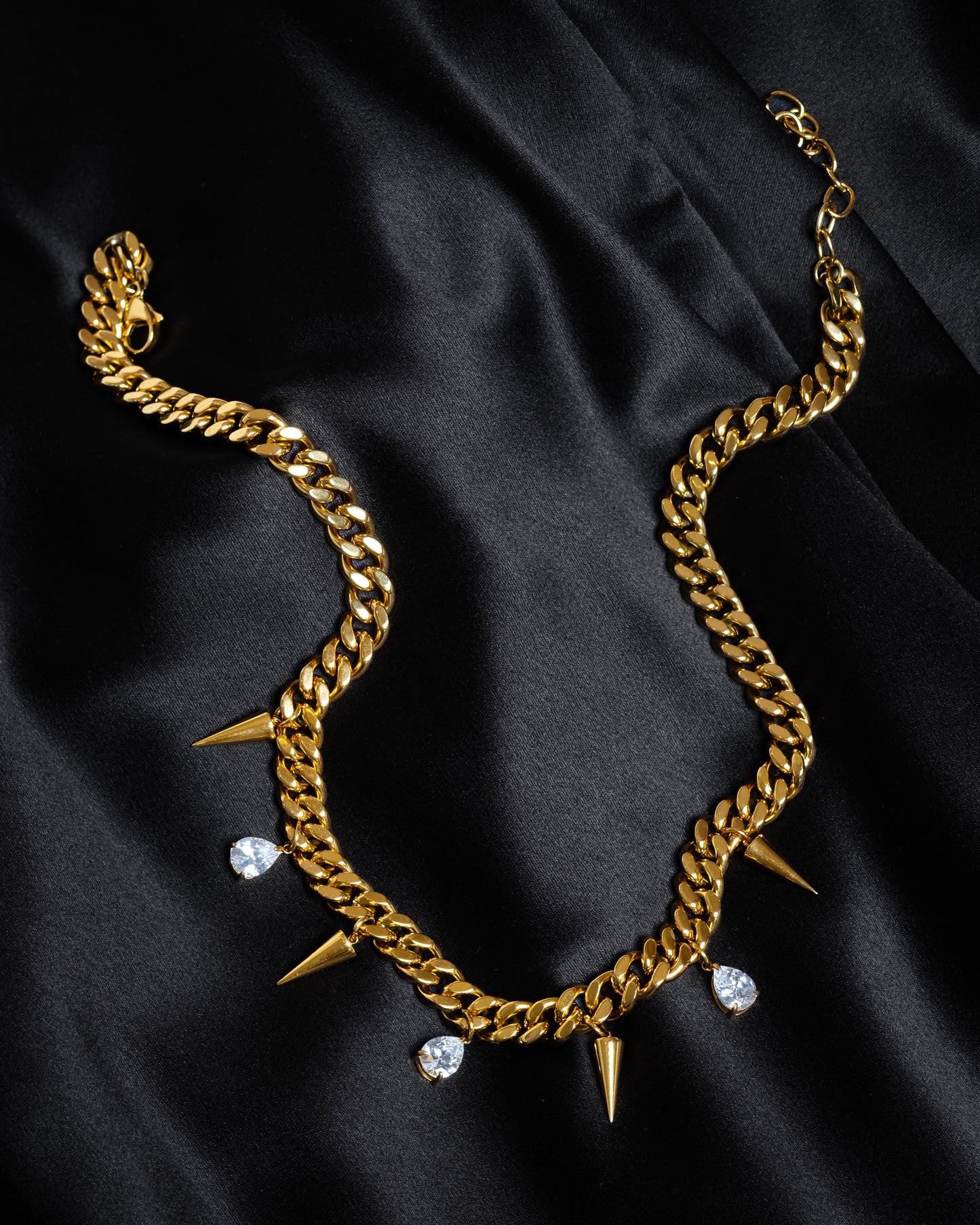 Detaljbild på guldkedja med nitar och vita kubiska zirkonia-stenar, liggande på svart siden.