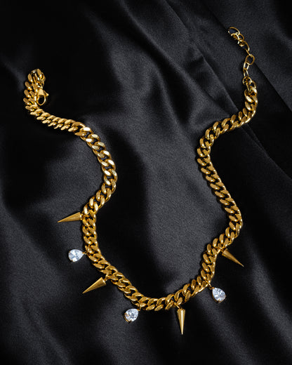 Detaljbild på guldkedja med nitar och vita kubiska zirkonia-stenar, liggande på svart siden.