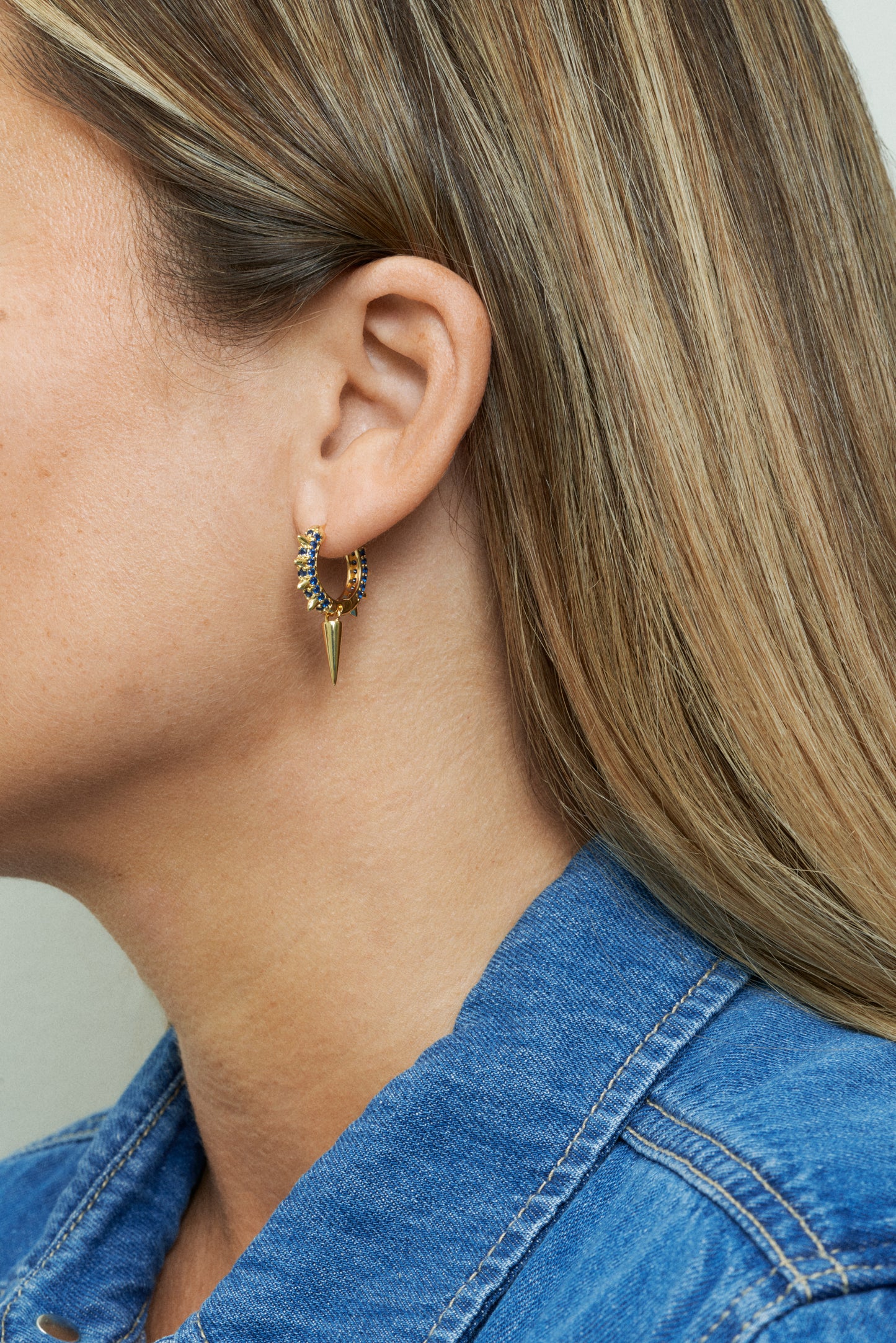 18k guldpläterade örhängen med blå stenar och nitdetaljer. Närbild på öra.