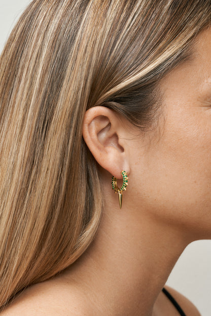Guld hoops med gröna zirkoner och spikdetaljer. Närbild på öra.