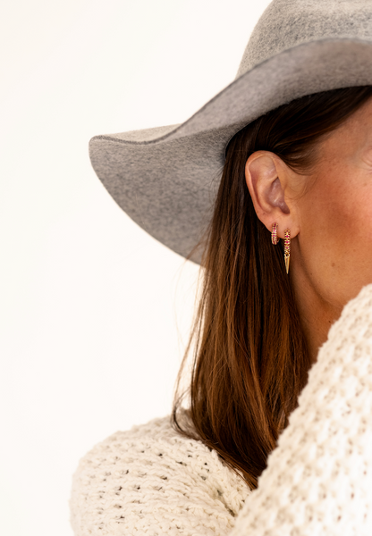 Röda zirkoner på guld hoops med spikdetaljer. Närbild på modell i vit kofta och grå hatt.